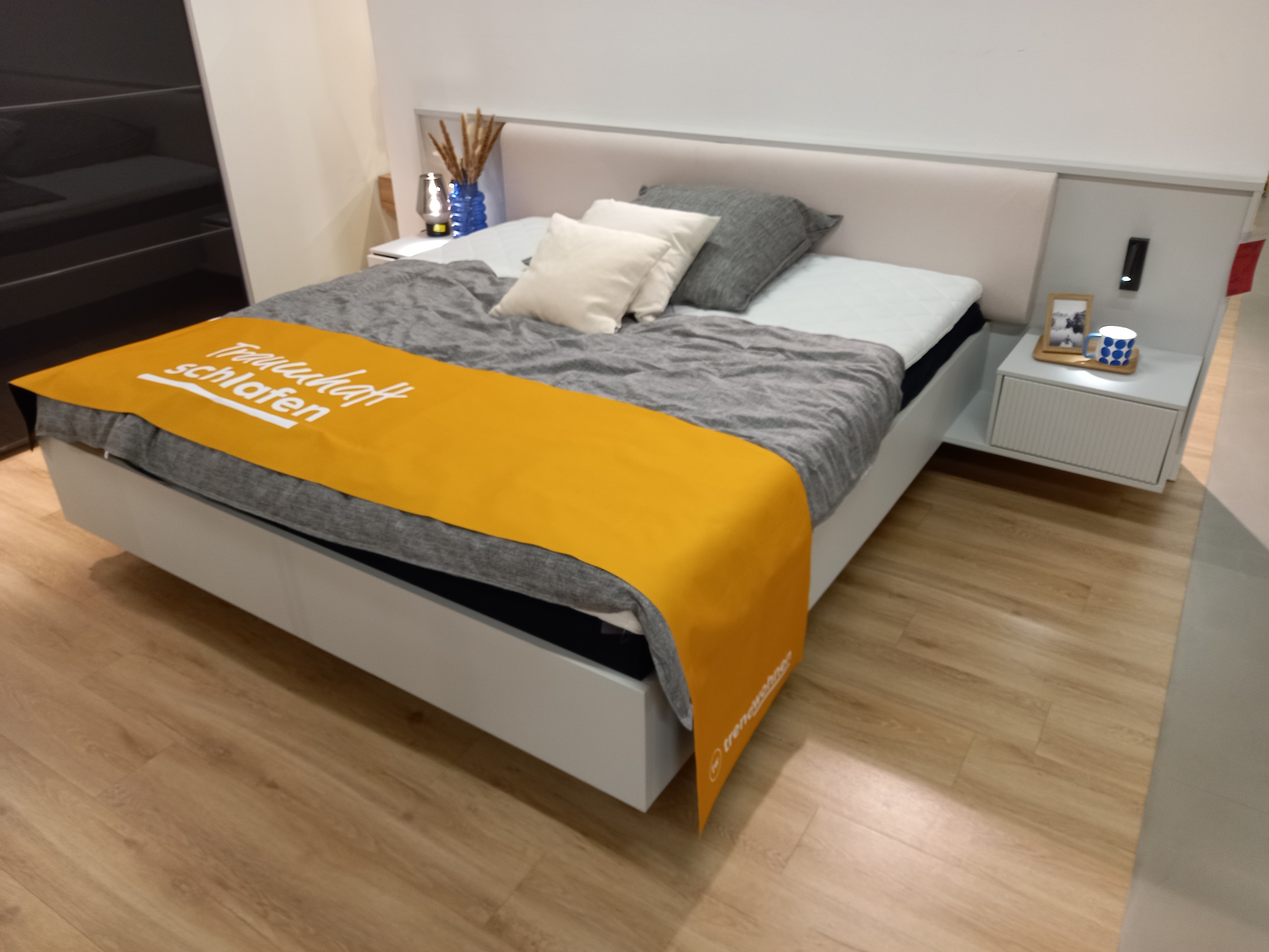 Bettanlage  in Farbe , Polsterbett, Bettgestell, Nachttisch, graue Tagesdecke, orangefarbener Bettrunner
